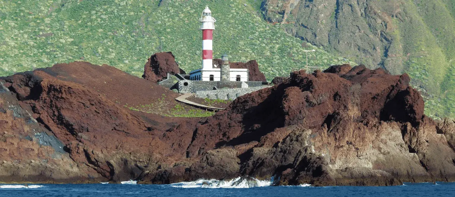 Red and white lighthouse on a volcanic rock, with buildings around it, blue sea and green mountain in the background.