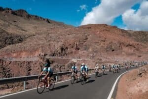 Gruppo di ciclisti in competizione su una strada accidentata, con paesaggio roccioso e montagne sullo sfondo.
