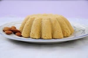 Torta dalla texture visibile, mandorle a sinistra, su piatto decorato, sfondo lilla chiaro.