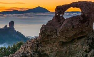 "Arco natural y roca en la cima, con montañas y nubes en el horizonte durante el atardecer."