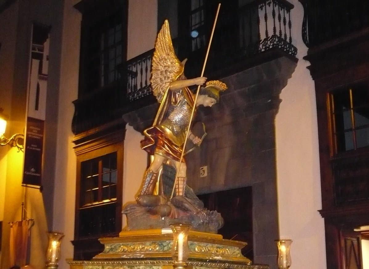 Estatua de una figura angelical, probablemente San Miguel, representada con una armadura dorada, alas y una lanza, de pie sobre un pedestal con un edificio histórico como telón de fondo.