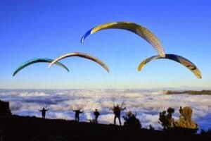 Parapendisti sopra una nuvola, con paracadute multicolore, in un paesaggio montano sotto un cielo azzurro.