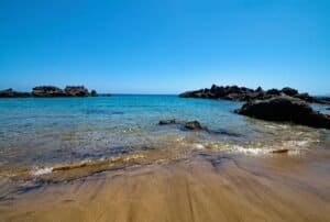 Playa con arena dorada, agua cristalina y rocas negras, cielo azul despejado.