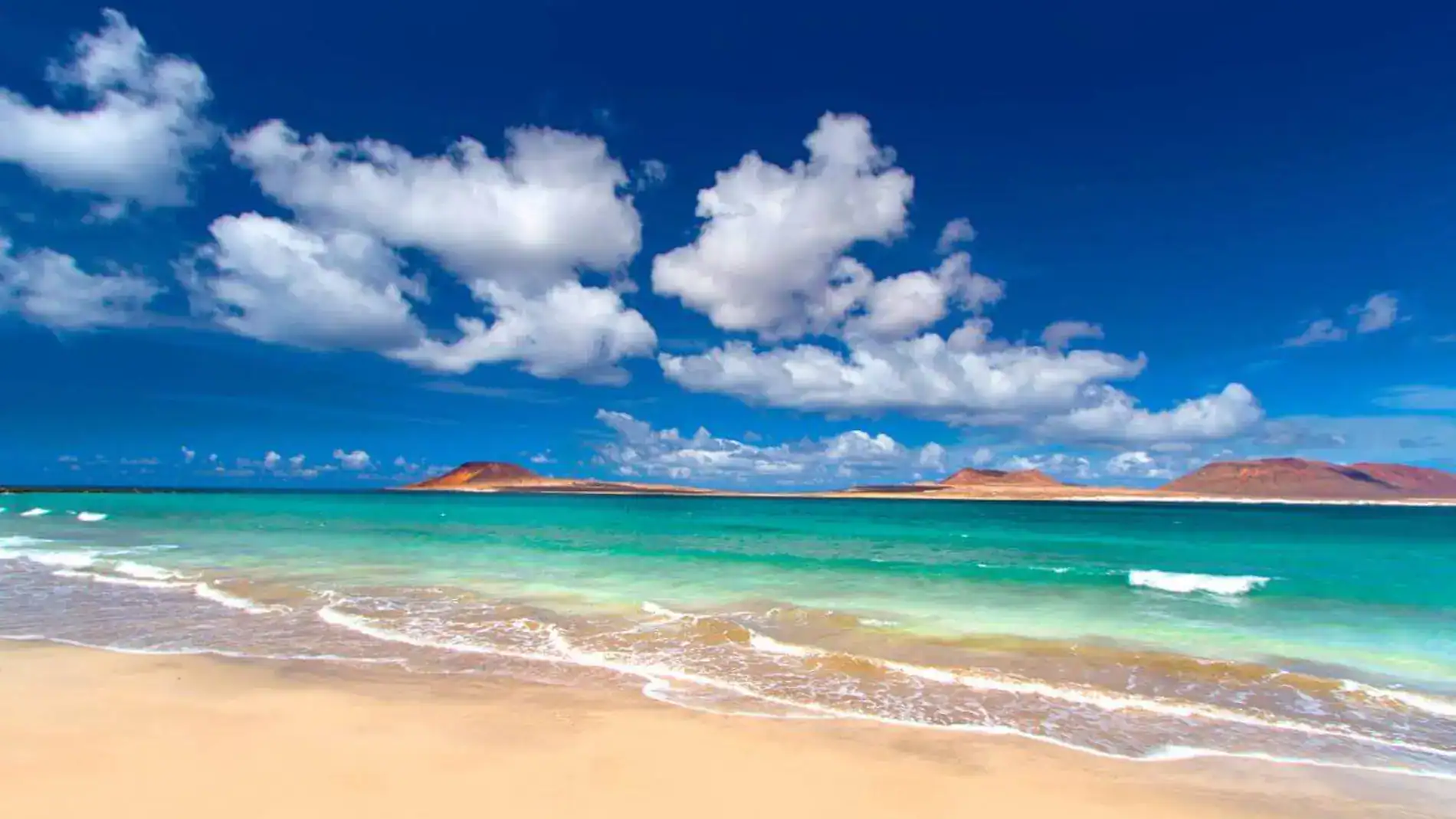 Playa con arena dorada, océano turquesa, nubes blancas, montañas rojas en el horizonte, cielo azul profundo.