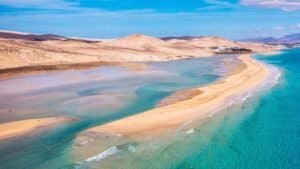 Playa con arena dorada, agua cristalina, montañas secas y cielo azul despejado.