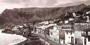 Pueblo costero con edificios blancos, montañas en el fondo, mar a la izquierda; paisaje histórico y rural.