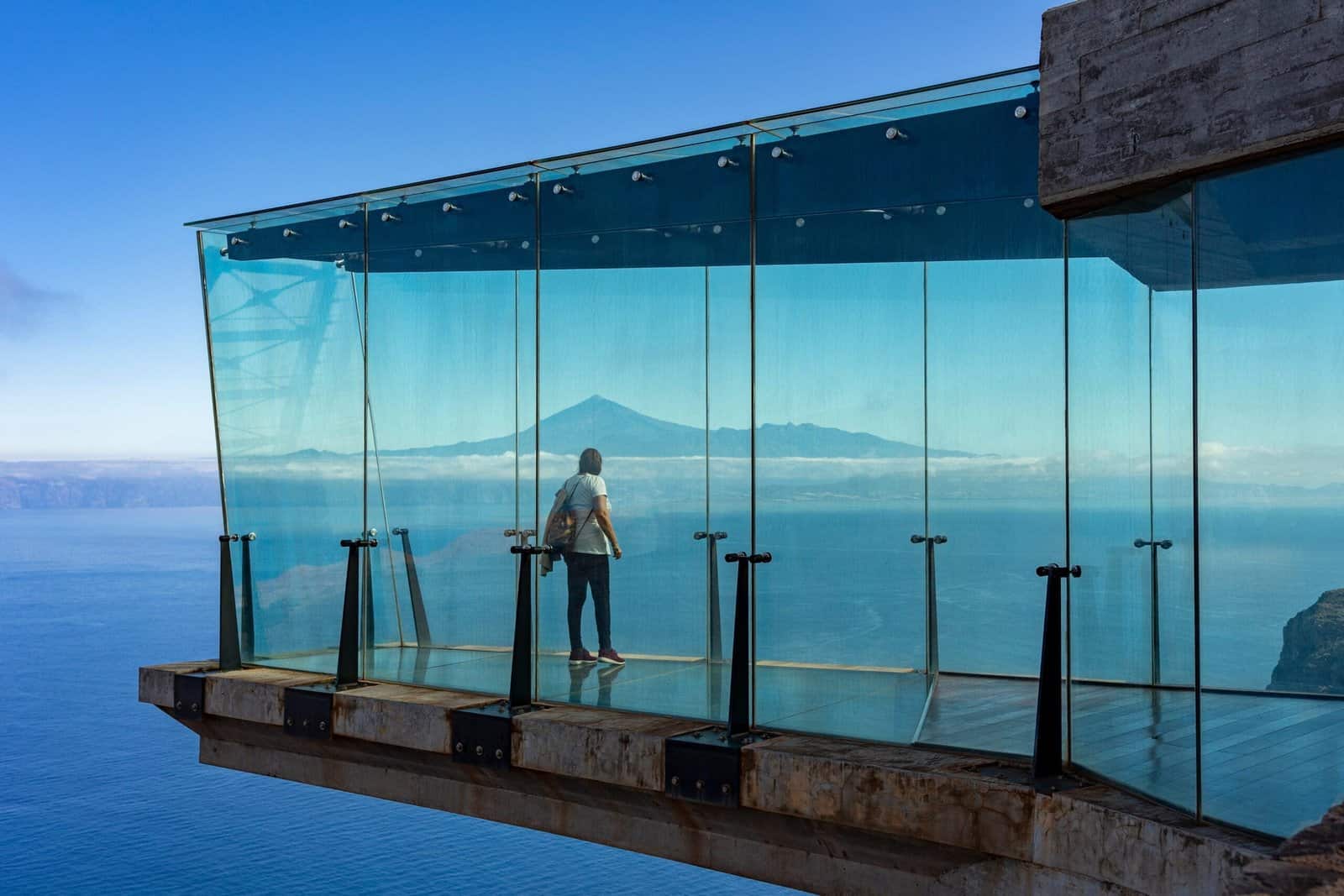 playas vírgenes. Una persona contempla el mar desde una plataforma de cristal suspendida sobre las olas, con un volcán visible en el horizonte bajo un cielo azul despejado.