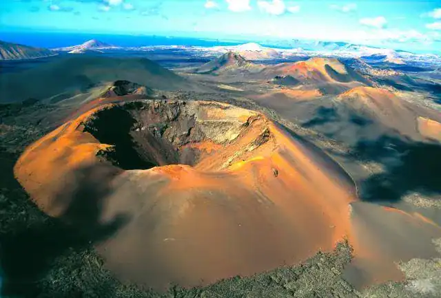 Volcán con cráter central, paisaje lunar con montañas circundantes, cielo azul y nubes dispersas.