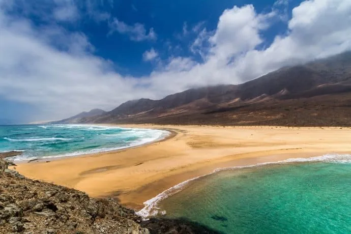 Beach with golden sand, turquoise sea, and mountains in the background, scattered clouds in the blue sky.