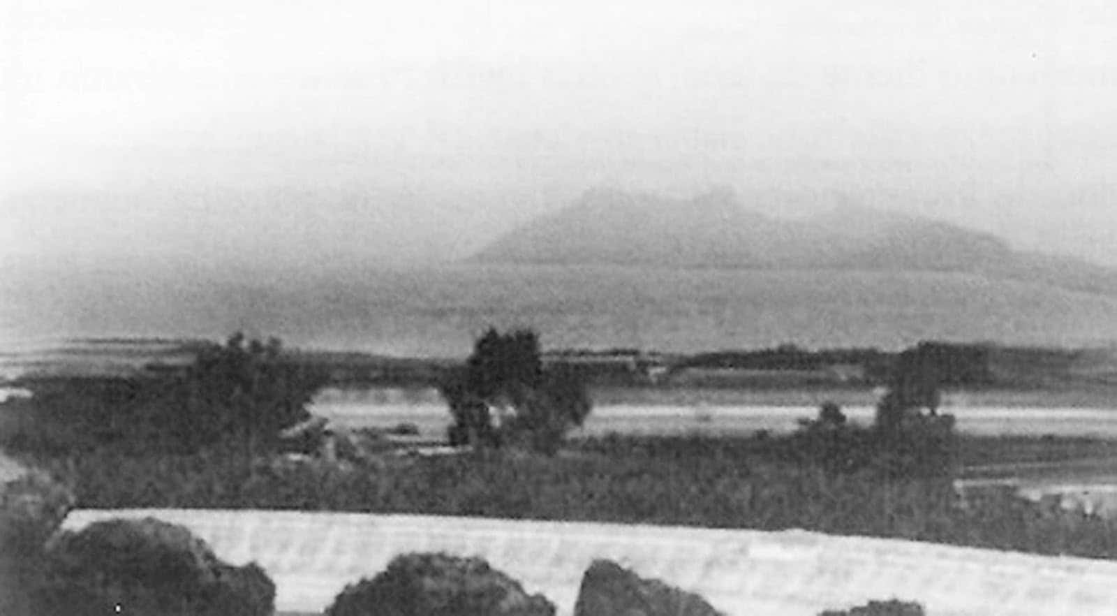 Montaña silueta en el horizonte, campos planos y bosque en primer plano; paisaje rural con atmósfera tranquila.