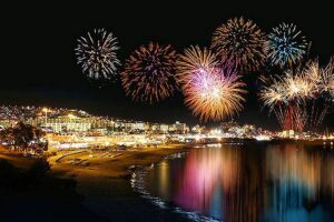 Fuegos artificiales sobre una ciudad costera iluminada, reflejos en el agua.