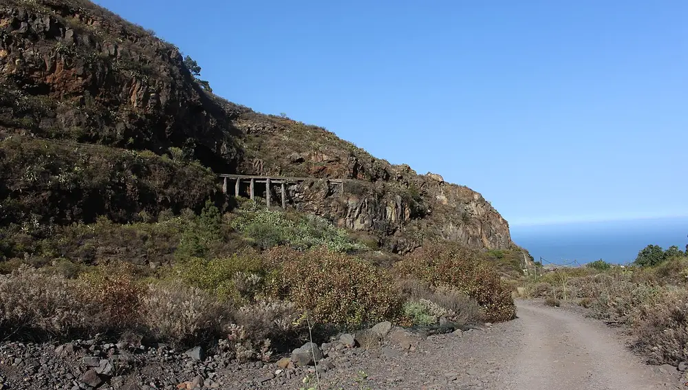 Camino de tierra que serpentea por una ladera rocosa con vegetación baja y seca, bajo un cielo azul despejado. En la pared de la montaña, se observa una estructura de madera antigua o en ruinas, posiblemente parte de un antiguo puente o pasarela. Al fondo, se divisa el mar en el horizonte, sugiriendo un entorno costero y natural, probablemente en una isla volcánica o zona mediterránea.