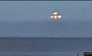 Objeto luminoso en el cielo nocturno o crepuscular, compuesto por cuatro luces brillantes agrupadas en una formación triangular o cuadrada difusa. El fondo es un cielo azul grisáceo con una línea horizontal que podría ser el horizonte marino o terrestre. La imagen está tomada a distancia y con baja resolución, lo que dificulta identificar el objeto con certeza —podría tratarse de un avión, dron, satélite, o simplemente un efecto óptico o de movimiento.