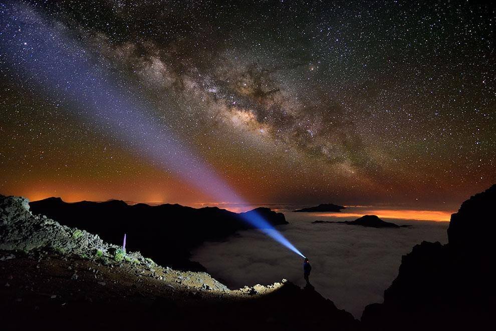 Una persona en silueta se alza sobre un acantilado volcánico, iluminando con una potente linterna el cielo estrellado mientras observa la Vía Láctea arqueándose majestuosamente sobre un mar de nubes. El horizonte brilla con un resplandor anaranjado, posiblemente de una ciudad lejana o el amanecer, contrastando con las sombras oscuras de las montañas y el suelo rocoso. La escena transmite soledad, asombro y conexión cósmica —ideal para representar la astrofotografía, la exploración nocturna o destinos como el Teide en Tenerife, donde el cielo es uno de los más limpios del mundo.