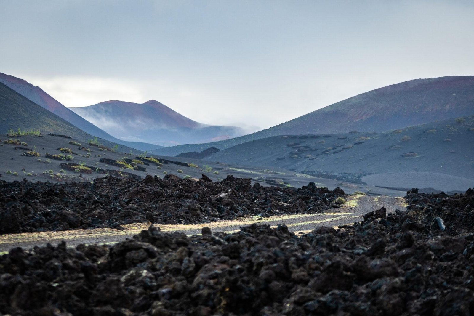 Paisaje volcánico desolado con suelo cubierto de rocas de lava oscuras y ásperas en primer plano. Al fondo, se extienden colinas y conos volcánicos de tonos rojizos y grises, parcialmente envueltos en niebla o bruma matutina. El cielo es claro pero tenue, sugiriendo la hora del amanecer o atardecer. Escasa vegetación verde brota entre las rocas, destacando la resiliencia de la vida en este entorno extremo. Ideal para representar paisajes lunares, parques naturales volcánicos como Timanfaya (Lanzarote) o zonas de alta actividad geológica.