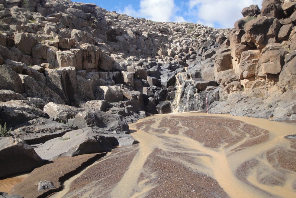 Pequeña cascada de agua turbia fluye entre formaciones rocosas volcánicas oscuras y angulosas, creando patrones intrincados en el lecho de arena y grava húmeda. El agua, de tono marrón amarillento, serpentea por el cauce antes de caer desde una altura modesta. El cielo azul con nubes blancas contrasta con la dureza del paisaje árido y erosionado, sugiriendo un entorno volcánico o semiárido, posiblemente tras una lluvia reciente. Ideal para representar erosión natural, geología volcánica o ecosistemas de arroyos efímeros.