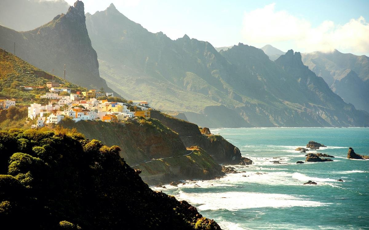 Vista panorámica de un pintoresco pueblo costero asentado en la ladera de una montaña escarpada, con casas blancas y de colores vivos apiladas sobre el acantilado. El mar turquesa rompe contra rocas negras en la base, mientras que al fondo se elevan imponentes picos montañosos cubiertos de vegetación verde. La luz del sol ilumina suavemente la escena, creando contrastes entre sombras y resplandores, y algunas nubes flotan en el cielo azul. Un paisaje típico de las islas Canarias, como Tenerife o La Gomera, que combina belleza natural, arquitectura tradicional y dramatismo geográfico.
