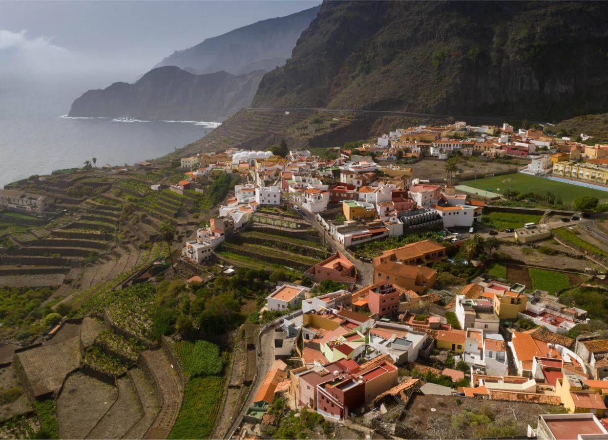 Vista aérea de un pintoresco pueblo costero enclavado entre montañas escarpadas y el mar. Las casas, de colores vivos —blancas, amarillas, naranjas y rosas— se apilan en terrazas que siguen el contorno de la ladera, rodeadas de campos agrícolas en terrazas y vegetación verde. Al fondo, acantilados rocosos caen hacia un océano grisáceo bajo un cielo parcialmente nublado. Se distingue una iglesia con cúpulas blancas, una cancha deportiva y caminos serpenteantes. La escena refleja la armonía entre la vida humana y el paisaje volcánico, típico de las islas Canarias, como La Gomera