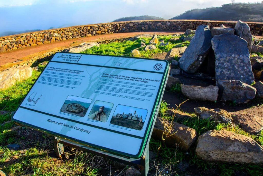 Panel informativo en primer plano del Mirador del Alto de Garajonay, con texto explicativo sobre el lugar, junto a un conjunto de grandes piedras megalíticas dispuestas en círculo sobre césped verde. Al fondo se observa un muro de piedra que rodea el mirador y un paisaje montañoso parcialmente cubierto por nubes.