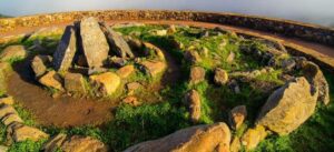 Círculo de piedras megalíticas en la cima de una montaña, con grandes rocas inclinadas en el centro rodeadas de hierba verde y un muro de piedra al fondo, bajo un cielo despejado.