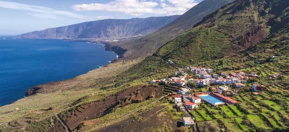 Vista aérea de un pintoresco pueblo costero enclavado entre montañas verdes y el mar azul profundo. Las casas blancas con techos rojos y naranjas se asientan en terrazas agrícolas que bajan hacia la costa volcánica. Al fondo, una cadena montañosa se extiende hasta el horizonte bajo un cielo azul con nubes dispersas. Se distingue una piscina comunitaria y caminos serpenteantes que conectan el pueblo con el entorno natural. La escena refleja la armonía entre la vida rural, la agricultura en terrazas y la belleza del paisaje insular —típico de las islas Canarias, como El Hierro.