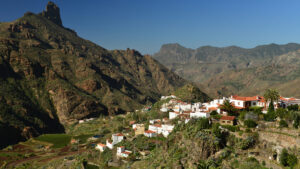 Vista panorámica de un pequeño pueblo blanco asentado en una ladera verde, rodeado por montañas escarpadas y rocosas bajo un cielo azul intenso. Las casas, con techos de teja roja y paredes brillantes, se distribuyen entre terrazas agrícolas y vegetación mediterránea, incluyendo palmeras y cactus. A la izquierda, un pico rocoso se alza majestuosamente sobre el valle. La luz solar resalta los contrastes entre sombras y colores, transmitiendo calma y autenticidad rural —escena típica de las islas Canarias, Gran Canaria.