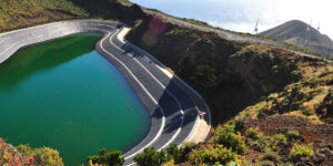 Depósito de agua de forma curva con revestimiento oscuro y agua de color verde intenso, situado junto a un acantilado. En la parte superior se observa una carretera y, al fondo, colinas con aerogeneradores y el océano bajo un cielo despejado.
