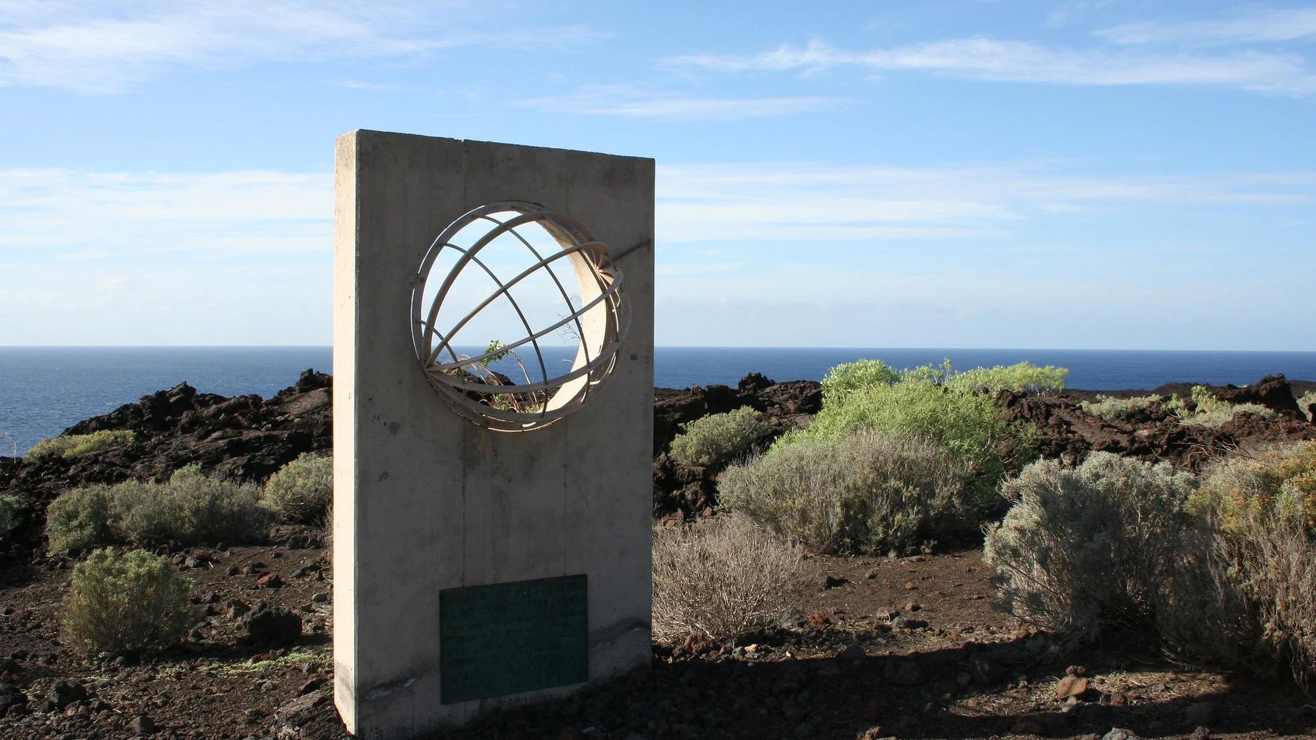 Monumento de hormigón situado en un paisaje volcánico junto al océano. La estructura presenta una esfera metálica atravesada por aros, simbolizando el globo terráqueo, y una placa en la base. Al fondo se observa el mar y un terreno oscuro de lava con vegetación baja, bajo un cielo despejado.