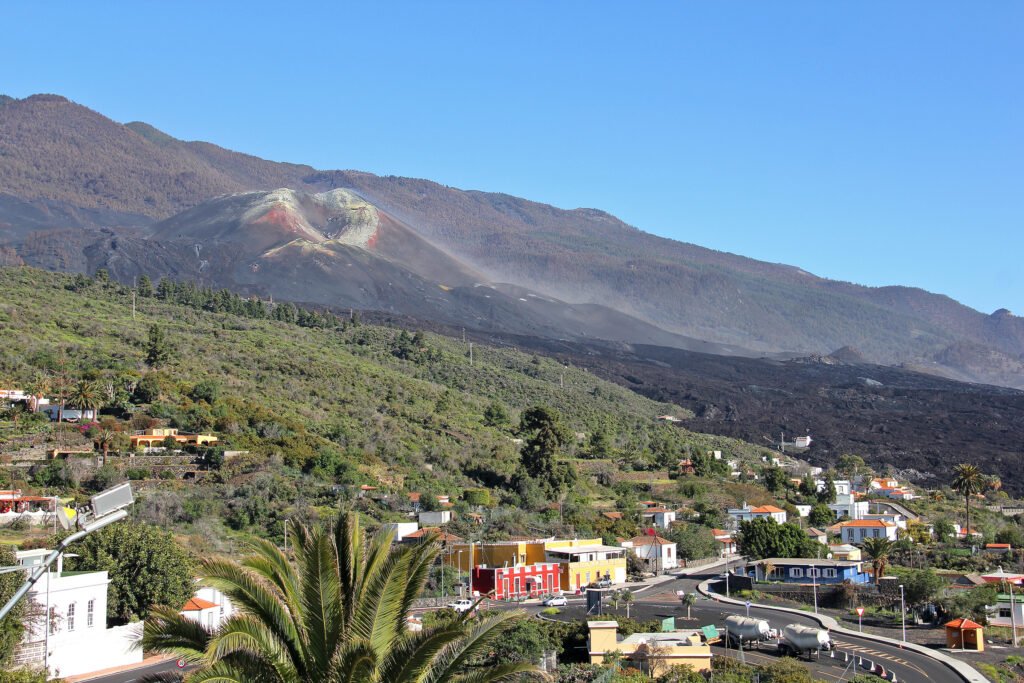 Vista de un paisaje volcánico con una montaña de tonos oscuros y rojizos en la cima, de la que se eleva una ligera nube de polvo o vapor. La ladera está cubierta por coladas de lava negra que descienden hacia una zona habitada con casas de colores, palmeras y carreteras que atraviesan el pueblo. El cielo está despejado y azul.
