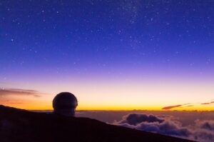 Observatorio astronómico con cúpula redonda en silueta sobre una montaña, bajo un cielo nocturno lleno de estrellas. En el horizonte se aprecia el resplandor del atardecer con tonos anaranjados y violetas, mientras un mar de nubes cubre la parte baja del paisaje.