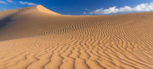 Primer plano de dunas de arena dorada con ondulaciones marcadas por el viento que crean patrones repetitivos en la superficie. Al fondo se eleva una duna más alta bajo un cielo azul con algunas nubes blancas, resaltando el contraste entre la textura de la arena y la claridad del día.