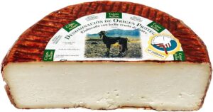 Mitad de una pieza de queso con corteza rojiza y marcada con un patrón acanalado. En la etiqueta se lee “Denominación de Origen Protegida” y muestra la imagen de una cabra, indicando que está elaborado con leche cruda de cabra. El interior del queso es compacto y de color blanco uniforme.