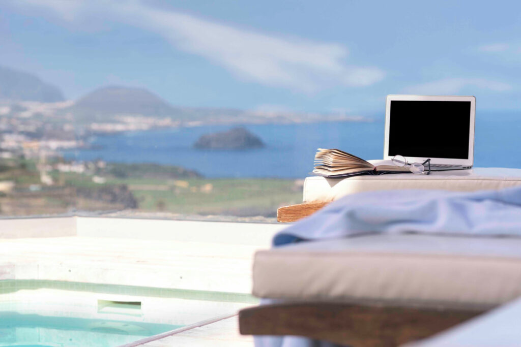 Laptop y libro junto a una piscina con vistas al mar y montañas en un destino costero