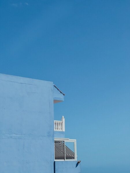 Vista minimalista de un edificio de paredes azul claro contra un cielo azul intenso. La construcción presenta detalles arquitectónicos blancos, incluyendo una balaustrada decorativa y un toldo de rayas blancas y negras en una ventana. Composición geométrica con el edificio ocupando el lado izquierdo del encuadre y el cielo despejado dominando la mayor parte de la imagen.
