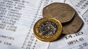 Monedas de libra esterlina británica sobre un ticket de compra de supermercado con un total de GBP 14.83. El recibo muestra diversos productos alimenticios como pasta, tomate, zanahoria, salchichas y otros artículos básicos con sus respectivos precios.