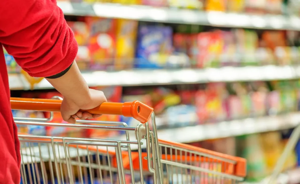 Primer plano de la mano de una persona con una camiseta roja empujando un carrito de compras con mango naranja en un supermercado. Las estanterías del fondo aparecen desenfocadas, mostrando productos coloridos en los pasillos del establecimiento.