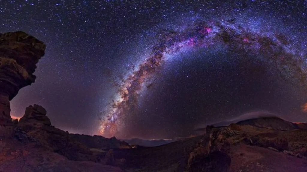 Paisaje nocturno con formaciones rocosas en primer plano y montañas al fondo, bajo un cielo espectacular donde la Vía Láctea forma un arco luminoso lleno de estrellas y tonalidades violáceas y azuladas. El horizonte muestra un leve resplandor cálido, contrastando con la oscuridad del terreno volcánico.
