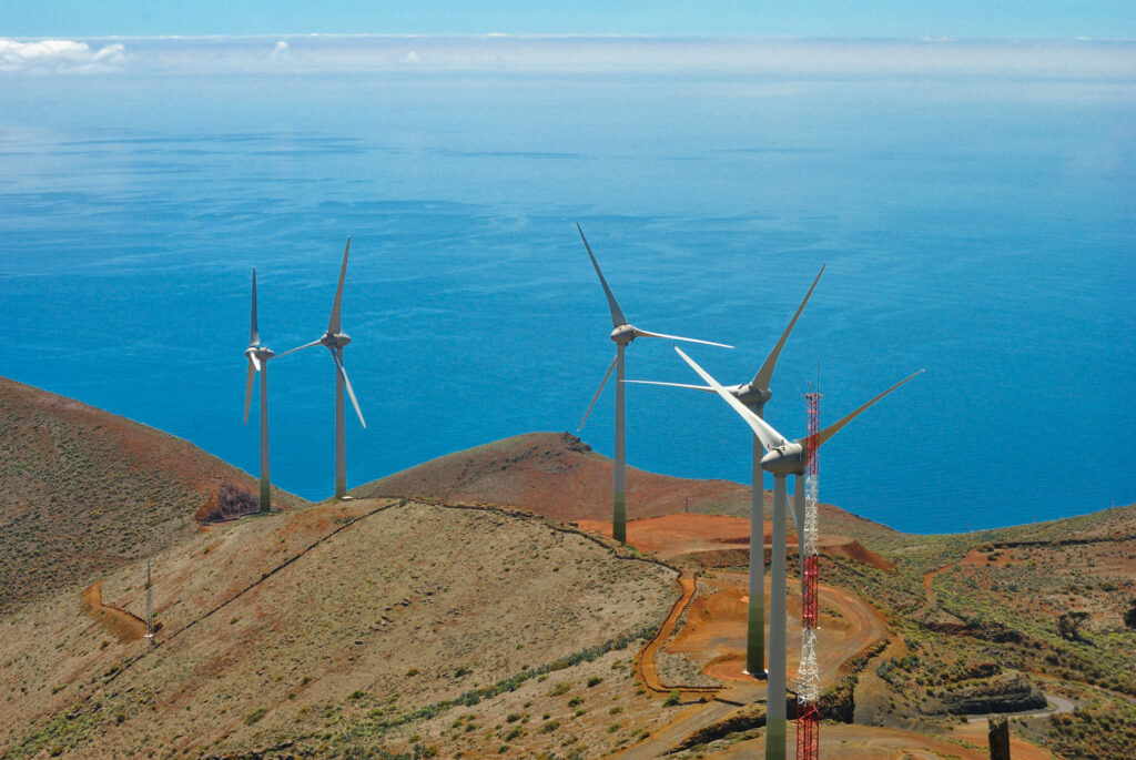 Conjunto de aerogeneradores situados sobre colinas áridas junto al océano. Las grandes turbinas blancas se alzan sobre el terreno rojizo y seco, conectadas por caminos de tierra, mientras el mar azul se extiende en el horizonte bajo un cielo despejado.
