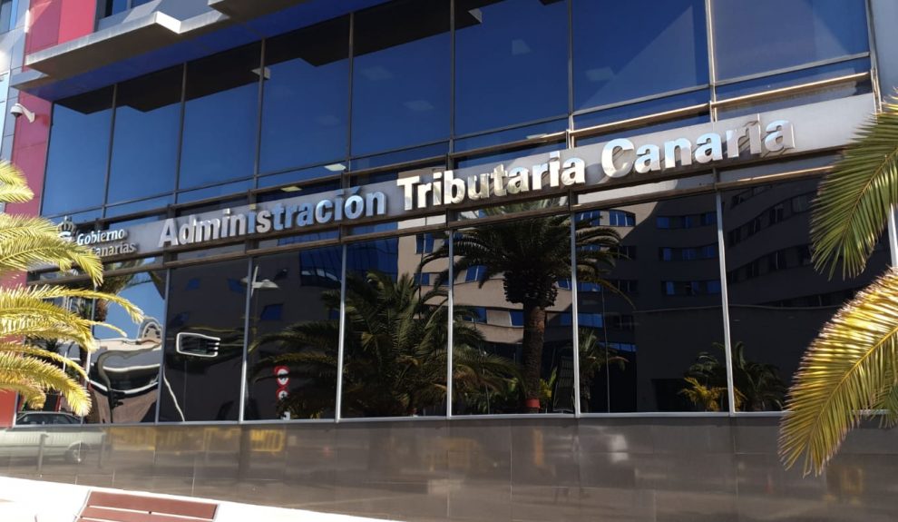 Fachada de un edificio moderno con grandes ventanales reflectantes y el rótulo “Administración Tributaria Canaria”, rodeado de palmeras.
