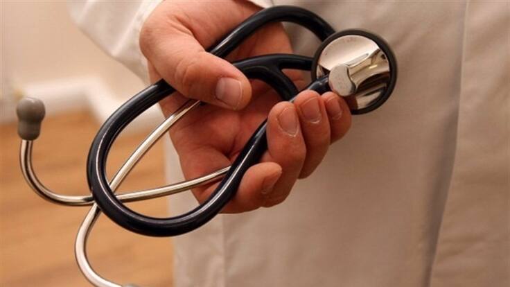 Primer plano de una mano sosteniendo un estetoscopio, con una bata médica al fondo, representando atención sanitaria o servicios de salud.
