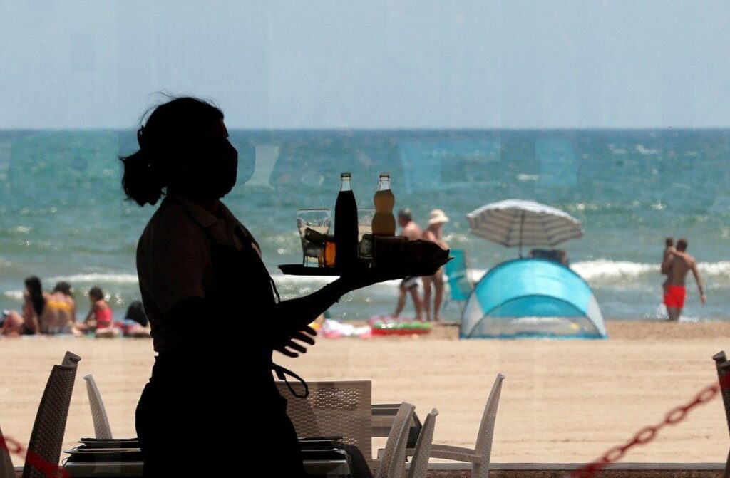 Trabajador con bandeja de bebidas en primer plano, playa con bañistas y sombrillas desenfocadas al fondo