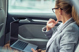 Mujer con gafas sentada en el asiento trasero de un coche, utilizando un ordenador portátil mientras mira hacia la ventana.