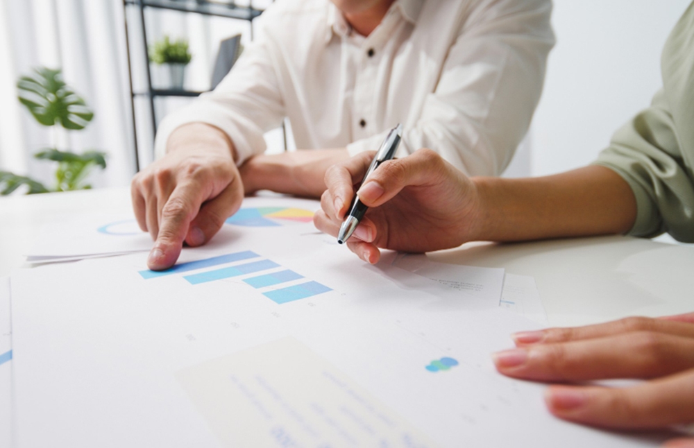 Dos personas analizando documentos con gráficos y datos, señalando y escribiendo sobre el papel durante una reunión de trabajo.