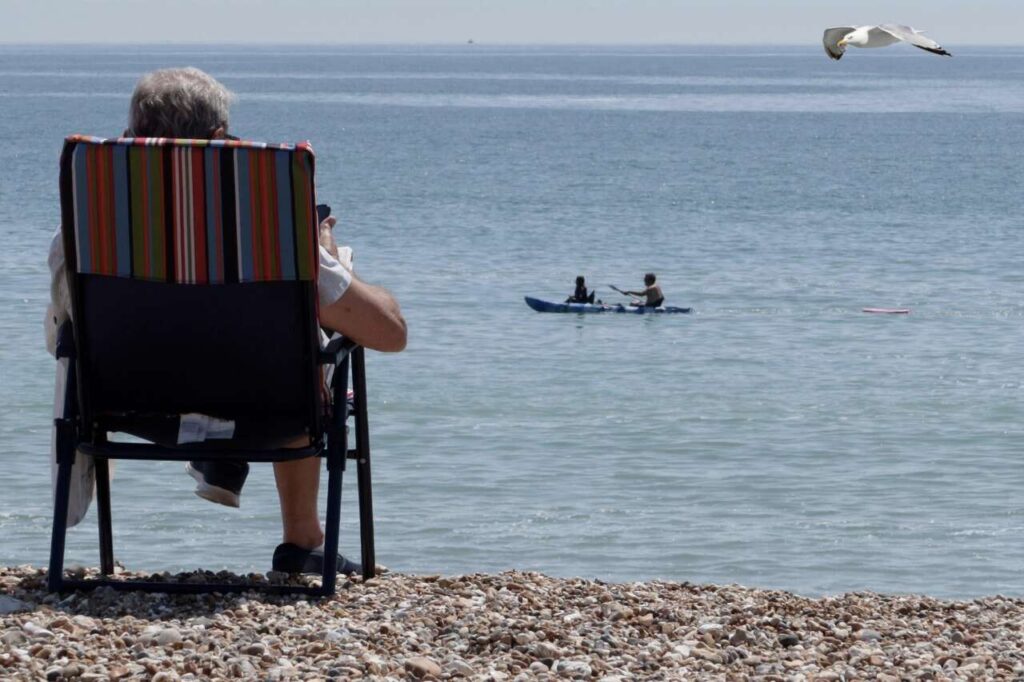Persona mayor sentada en una silla de playa sobre la orilla de piedras, mirando el mar tranquilo mientras, a lo lejos, dos personas navegan en kayak y una gaviota vuela sobre el agua.
