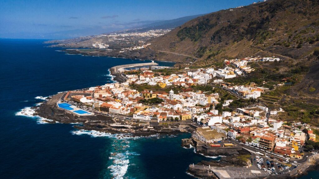 Vista aérea de un pueblo costero con casas blancas y tejados rojizos junto al mar, rodeado de acantilados y montañas, con olas rompiendo en la costa.
