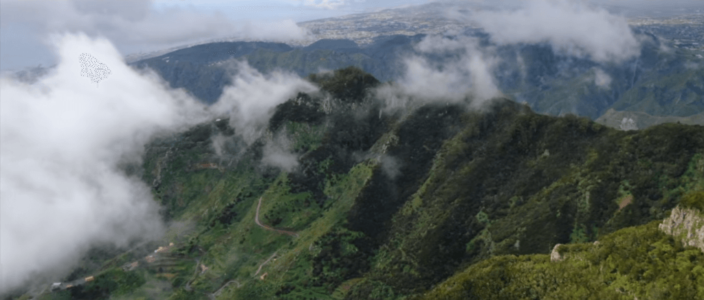 Paisaje montañoso cubierto de vegetación verde, con nubes bajas rodeando las cumbres y un camino serpenteante visible en la ladera.
