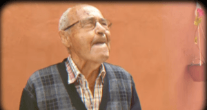 Hombre mayor con gafas, de pie frente a una pared anaranjada, mirando ligeramente hacia arriba con expresión pensativa. Lleva una camisa a cuadros y un suéter oscuro con patrón.