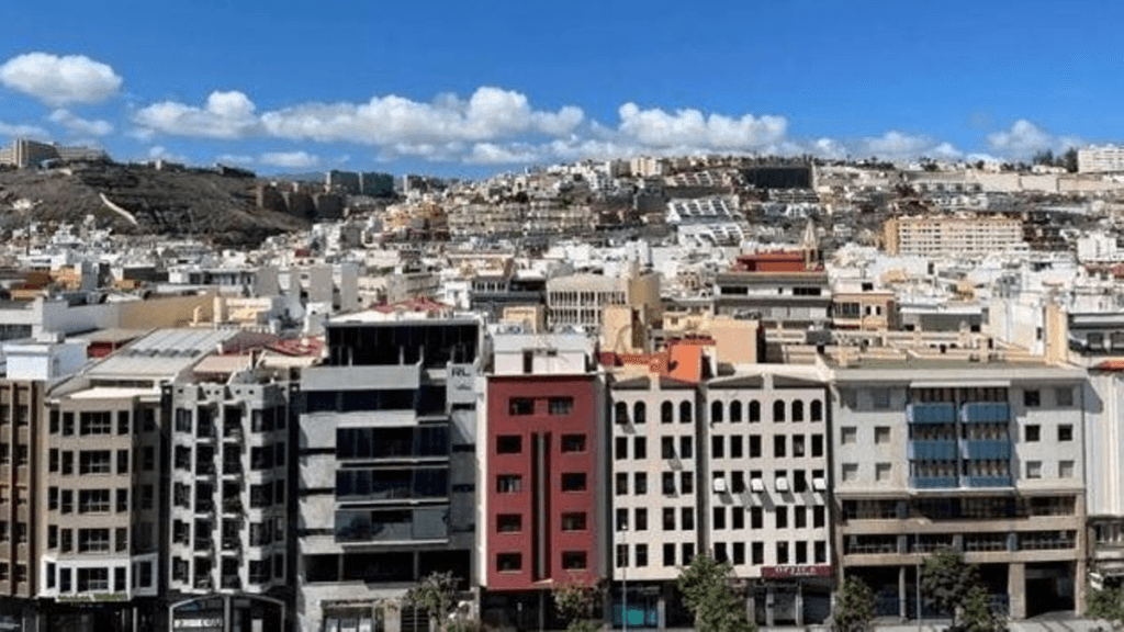 Vista panorámica de una ciudad con numerosos edificios residenciales y comerciales de varios pisos, extendidos por una zona urbana con colinas al fondo bajo un cielo parcialmente nublado.