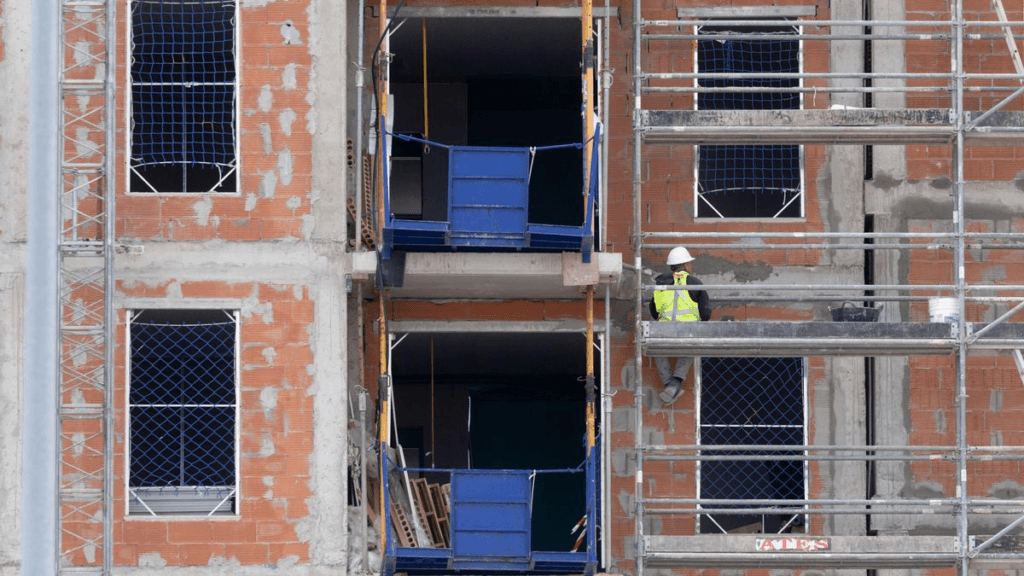 Trabajador de la construcción con casco y chaleco reflectante sobre un andamio, junto a un edificio en obra con paredes de ladrillo, ventanas sin terminar y estructuras de soporte.