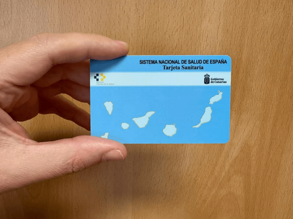 Mano sosteniendo una tarjeta sanitaria del Sistema Nacional de Salud de España, con diseño azul y un mapa de las Islas Canarias, sobre un fondo de madera.
